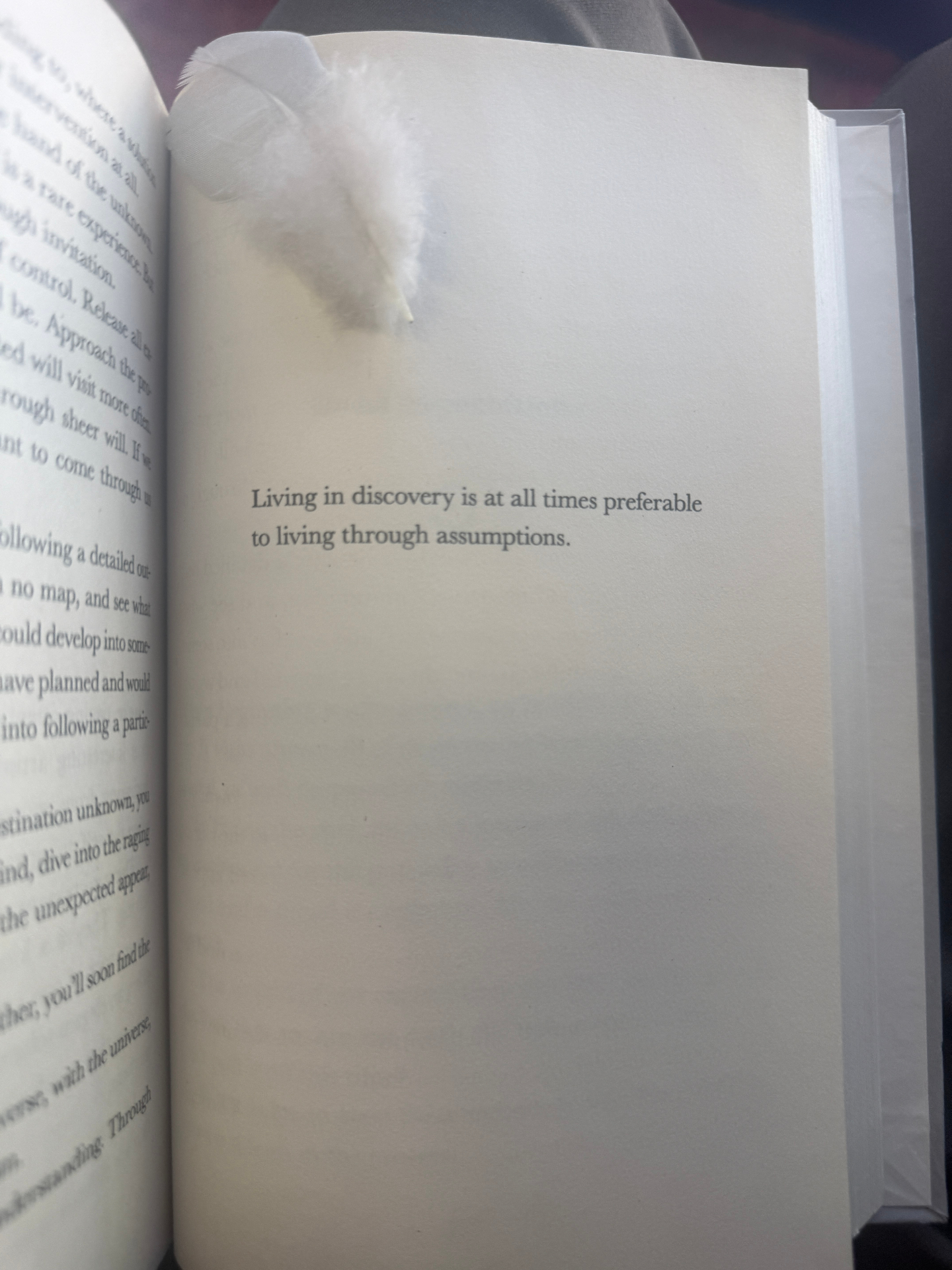 An open book with a white feather resting on the page, showing the passage 'Living in discovery is at all times preferable to living through assumptions.'